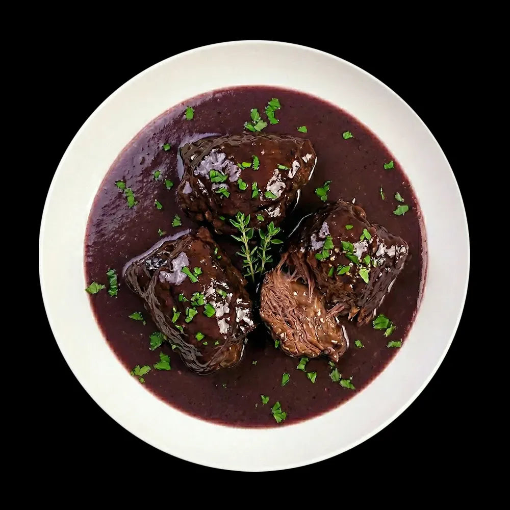 Servierfertige, zart geschmorte Rinderbäckchen in einer tiefroten, glänzenden Rotwein-Reduktion. Die butterweichen Fleischstücke sind auf einem weißen Teller mit frischen Kräutern garniert – ein Gourmet-Klassiker aus der Mainzer Küche der Metzgerei Walz.