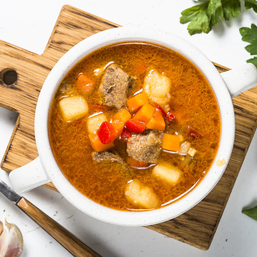 Kräftige Gulaschsuppe mit zarten Rindfleischwürfeln, Kartoffeln und Paprika in aromatischer, rötlich-brauner Brühe, serviert in einer weißen Schale auf einem Holzbrett.