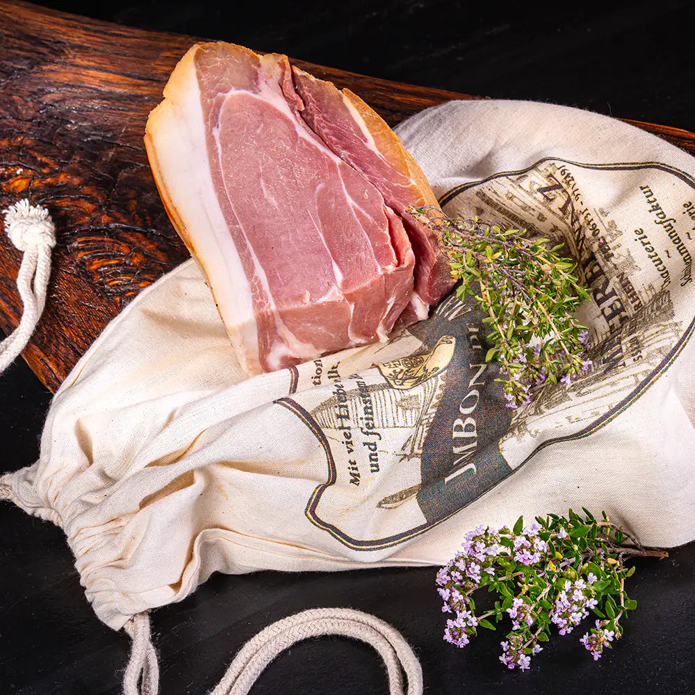 Mainzer Schinkenstück aus der Metzgerei Walz auf rustikalem Holzbrett mit Kräutern und traditionellem Stoffbeutel.