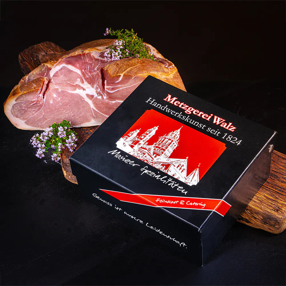 Verpackter Mainzer Spezialitäten-Schinken der Metzgerei Walz mit frischem Schinkenstück auf Holzbrett und Kräutern.